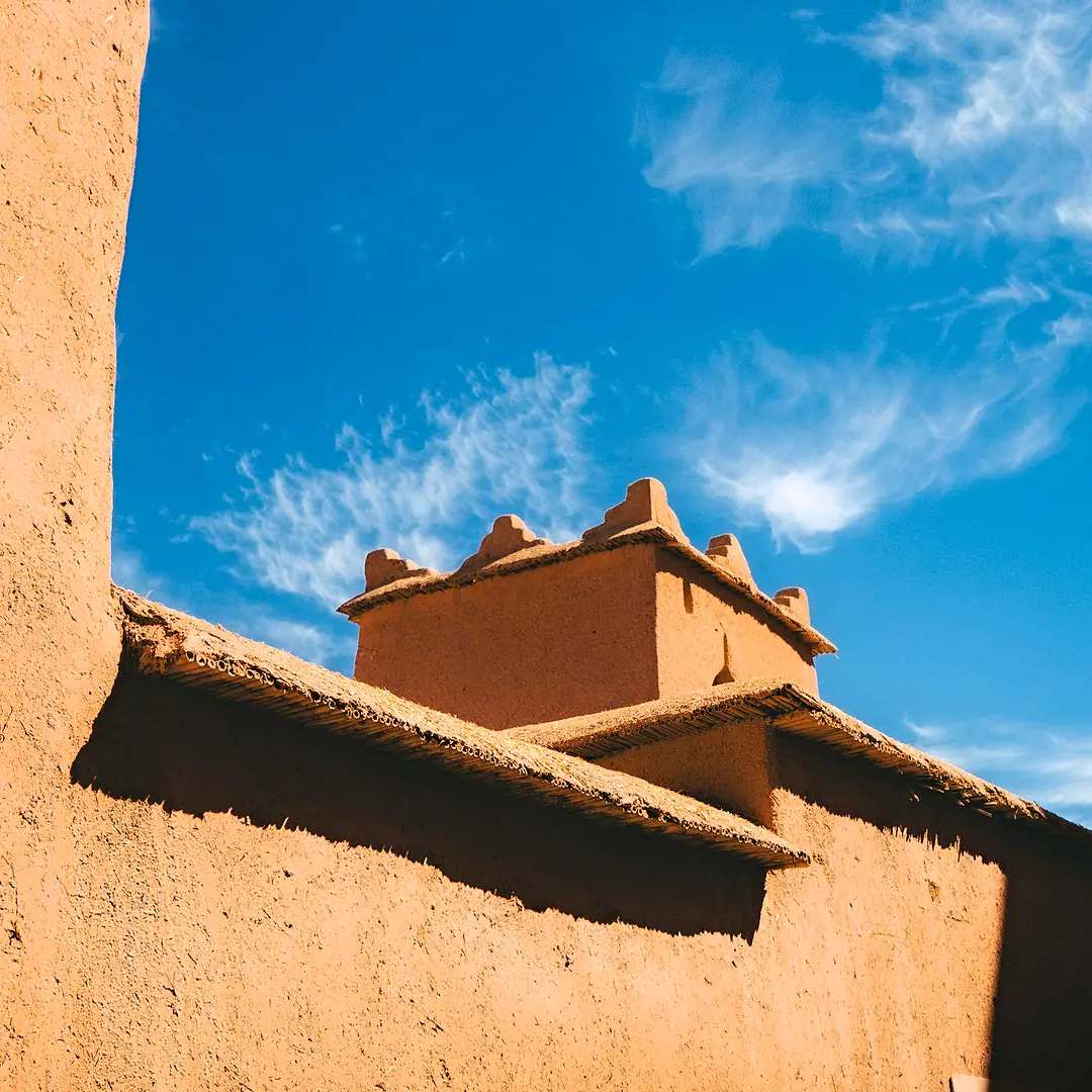 The rammed-earth walls and crenellated tower of a kasbah against a blue sky with high cloud