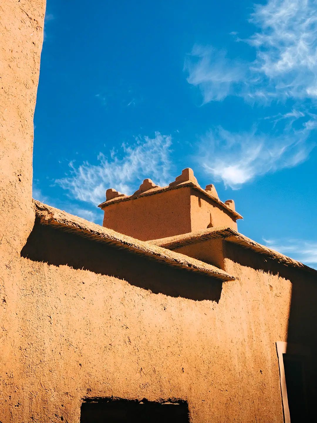 The rammed-earth walls and crenellated tower of a kasbah against a blue sky with high cloud