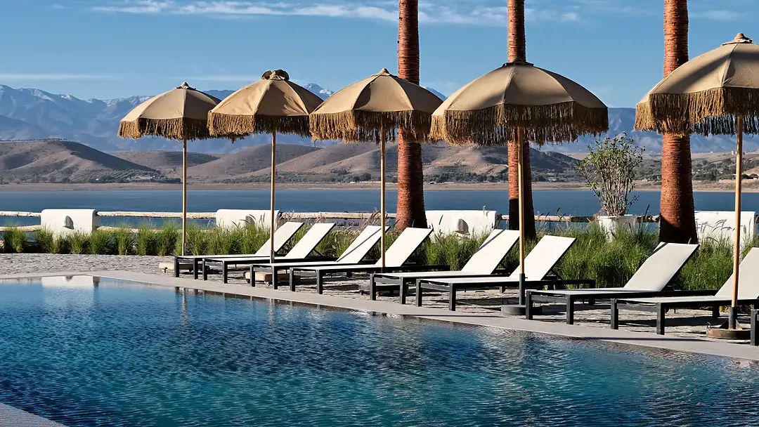 A resort pool lined with palms, fringed parasols and loungers, the Atlas mountains beyond