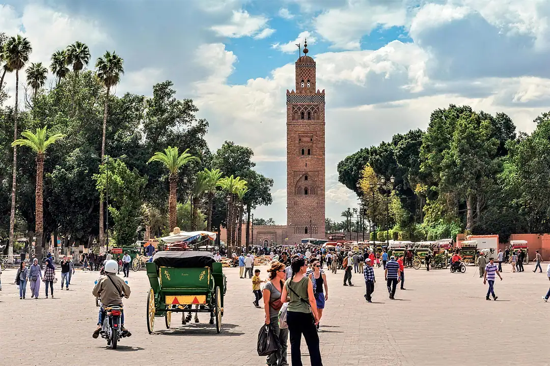 The Koutoubia minaret in Marrakech above a busy square, palms, a green horse-drawn carriage and visitors crossing the paving