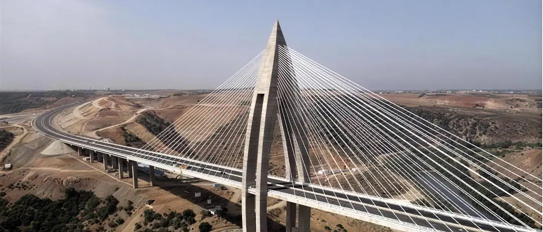 A cable-stayed motorway bridge from the air, its arched concrete pylon rising above a dry valley and the deck curving away across the hills