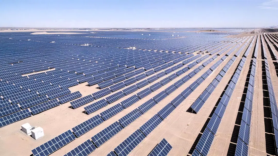 A photovoltaic plant from the air, long rows of solar panels across pale desert ground, a small white technical building in the foreground
