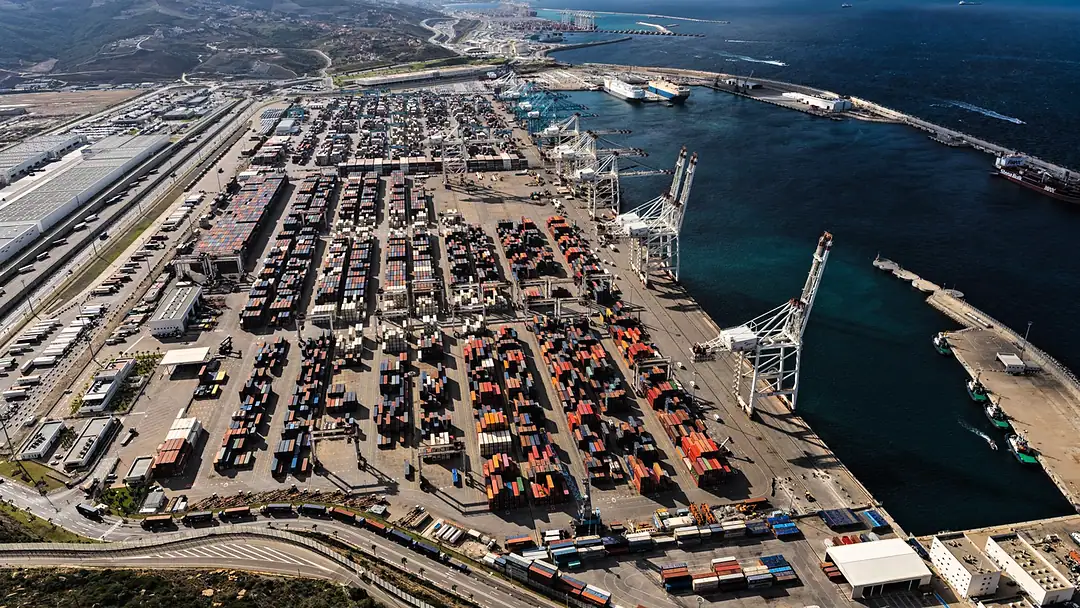 A container port and its logistics zone from the air, cranes along the quay, stacked containers and warehouses, hills beyond