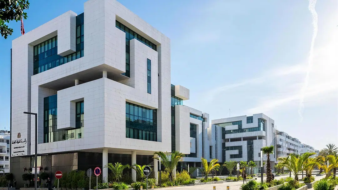 A white contemporary institutional building with deep-set windows and glass, palms and a wide pavement in front, under a blue sky
