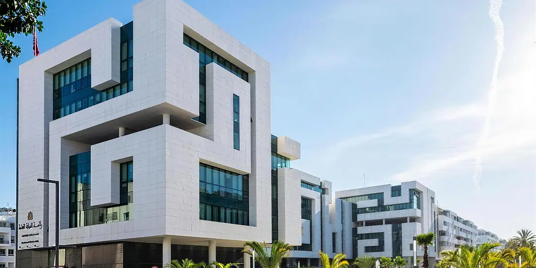 A white contemporary institutional building with deep-set windows and glass, palms and a wide pavement in front, under a blue sky