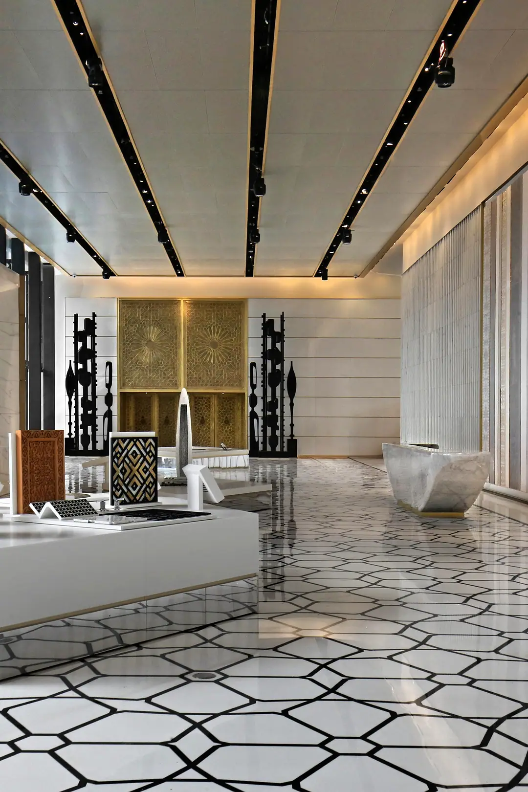 An institutional lobby with a patterned marble floor, a white display table in the foreground, a gilded carved screen and dark sculpted totems on the far wall, lines of light across the ceiling