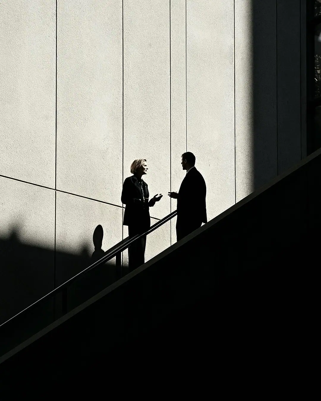 Two silhouetted figures talking on a staircase against a sunlit concrete wall